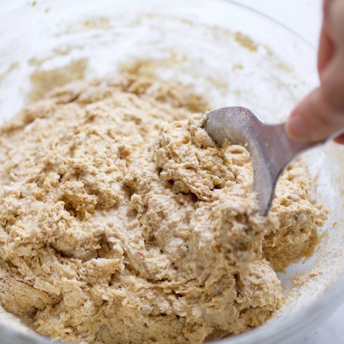 Whole-grain einkorn flatbread dough being mixed in a glass bowl with a metal dough scraper