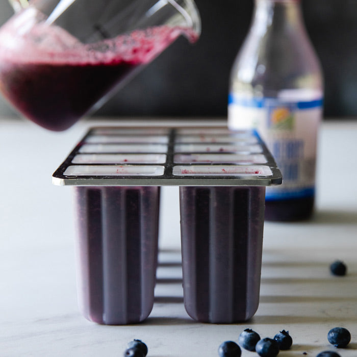 Pitcher pouring blueberry fruit nectar into popsicle molds on a marble countertop with scattered blueberries