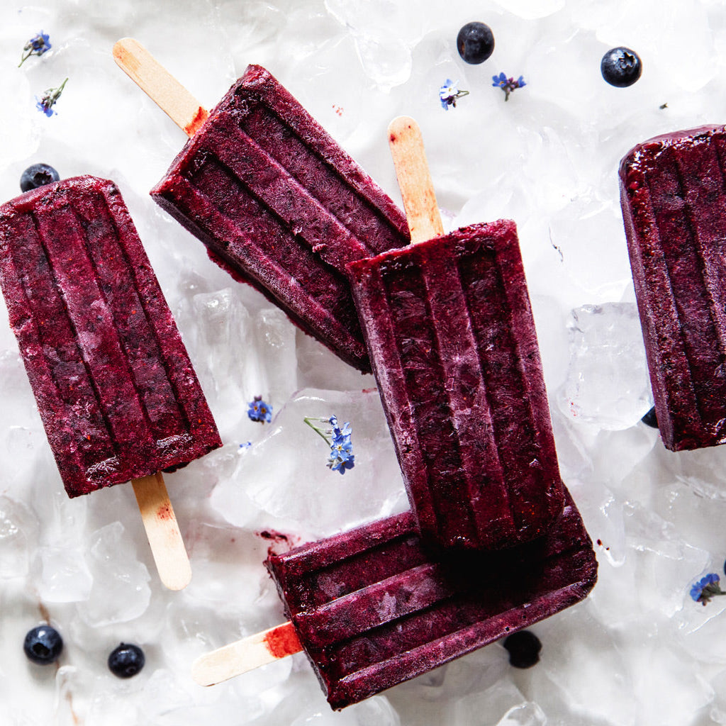 Three wild berry popsicles made from organic fruit spread on crushed ice with scattered blueberries and small blue flowers