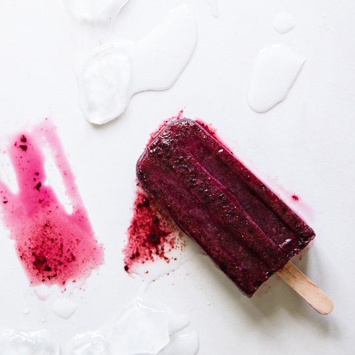 Top-down view of a deep purple organic fruit nectar popsicle on a white surface showing berry flecks, melting juices, and a wooden stick