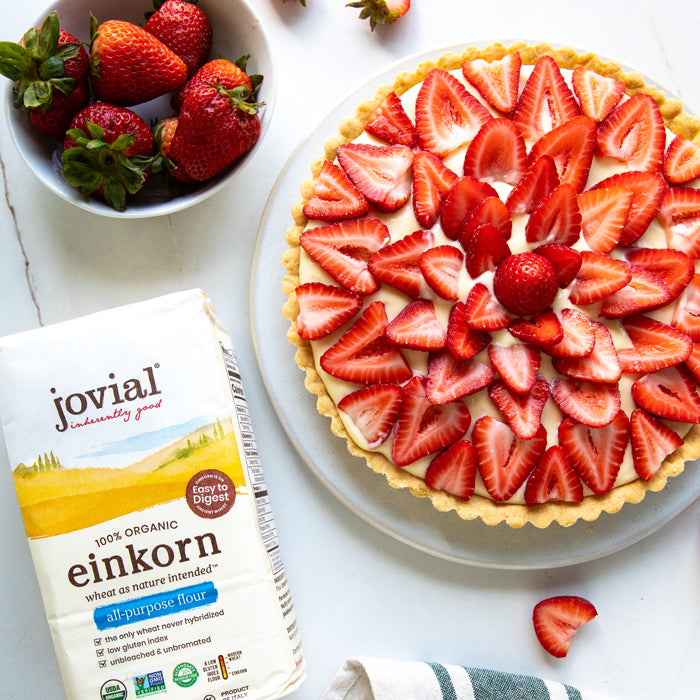 Top-down view of a strawberry fruit tart on a plate with a bag of Jovial organic einkorn flour and a bowl of strawberries