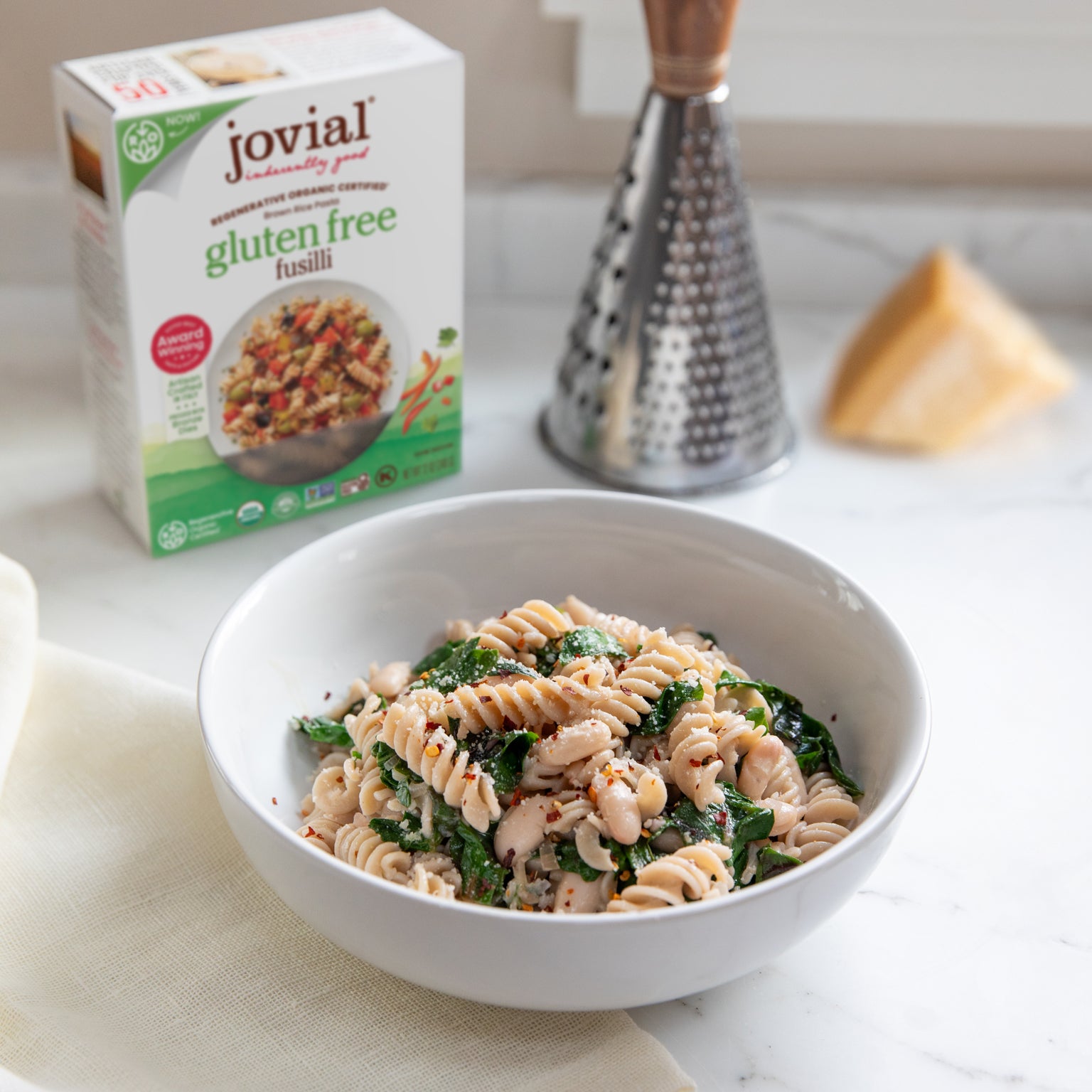 Bowl of fusilli with Swiss chard and cannellini beans with a Jovial gluten free fusilli box and cheese grater behind