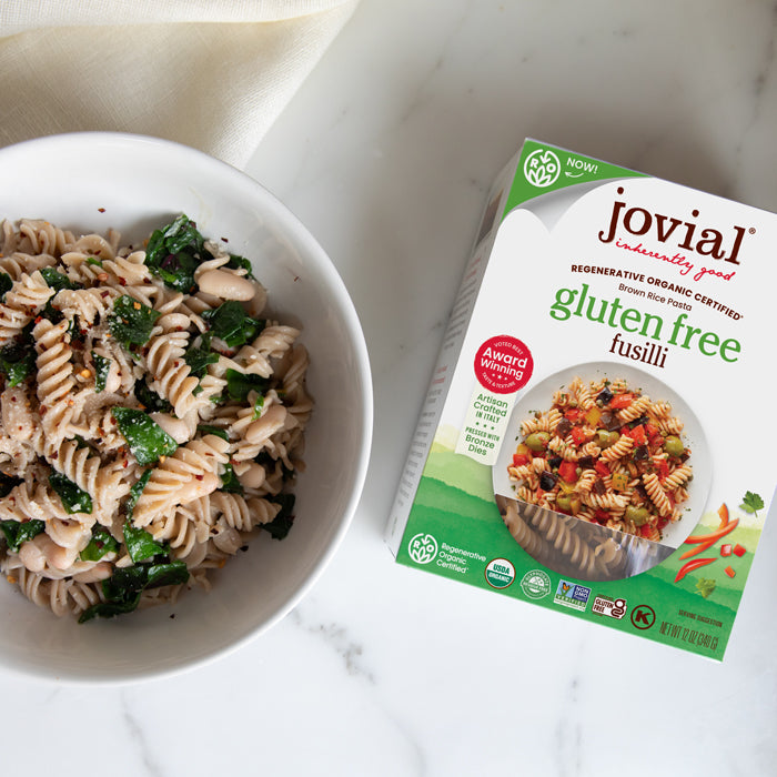 Top-down view of Jovial gluten free fusilli box beside a bowl of cooked fusilli with Swiss chard on a marble surface