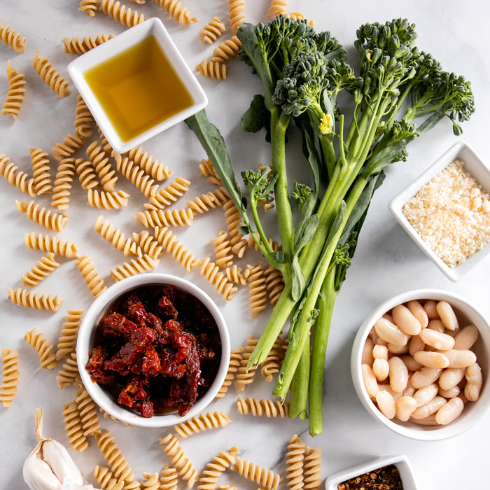 Fusilli ingredients arranged on marble: einkorn fusilli, rapini, cannellini beans, sun-dried tomatoes, olive oil, cheese