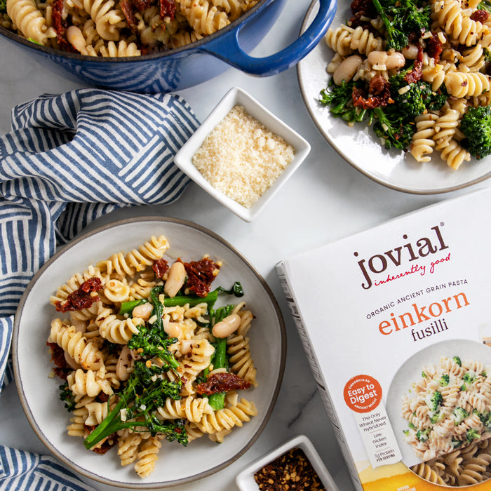 Bowl of fusilli with rapini, cannellini beans, and sun-dried tomatoes beside a box of Jovial Einkorn fusilli on marble
