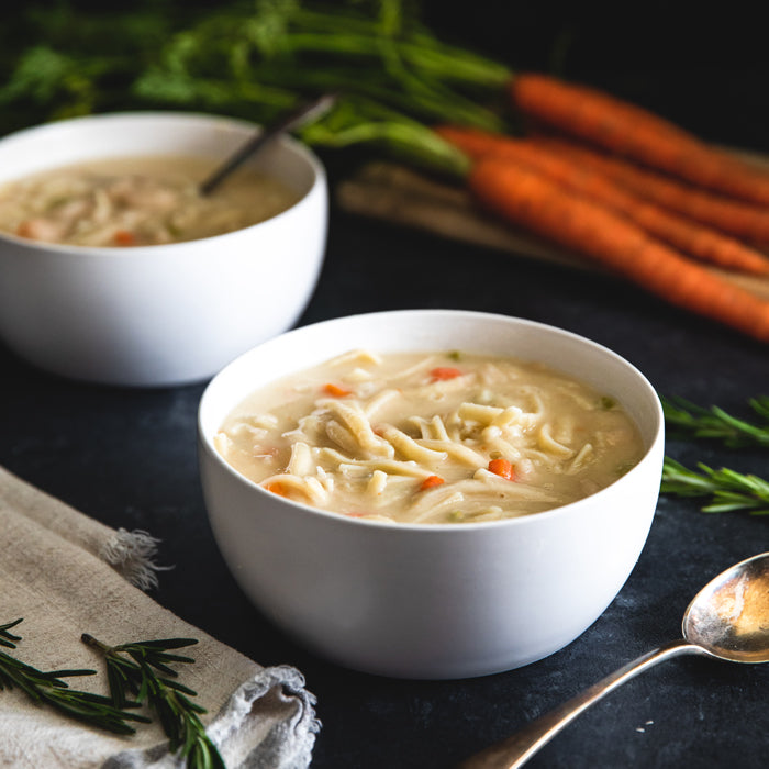 Two bowls of creamy cannellini bean soup with gluten-free linguine on a dark table, carrots and rosemary in the background