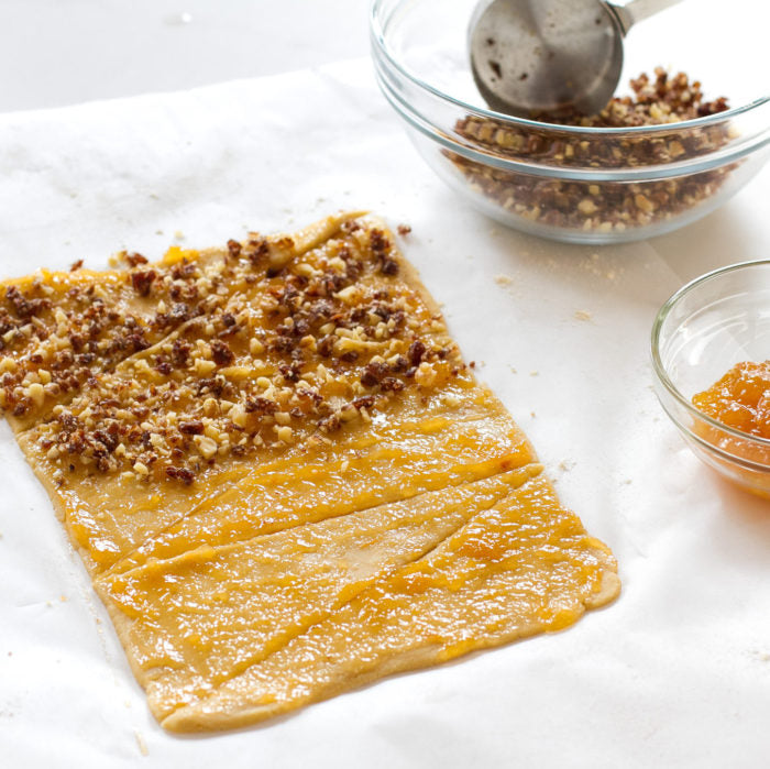 Einkorn rugelach dough spread with apricot jam and sprinkled with chopped nuts on parchment paper, glass bowls nearby