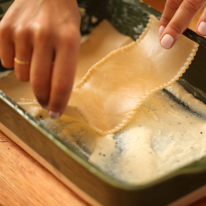 Hands assembling gluten-free lasagna, placing a lasagna sheet over white cheese sauce in a rectangular baking dish