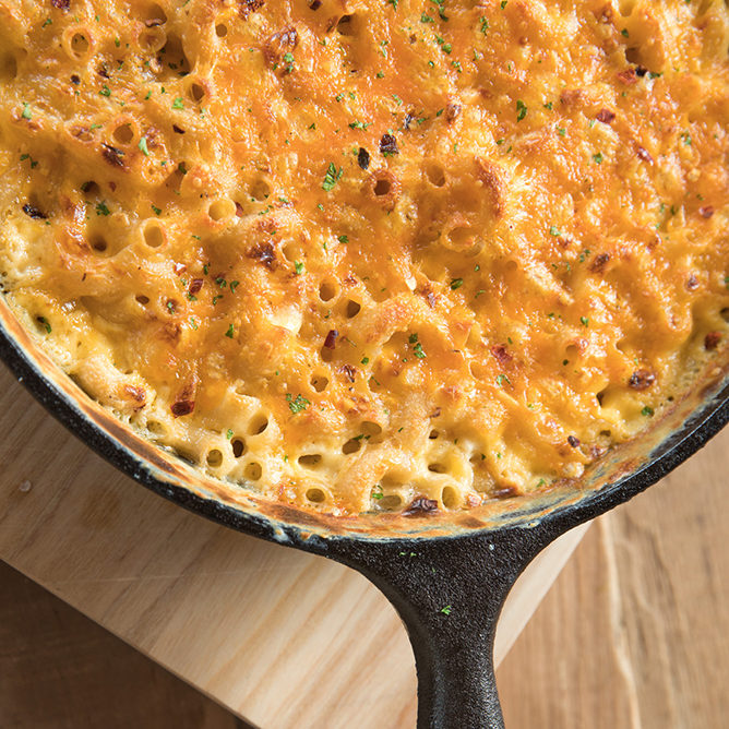 Gluten-free mac and cheese broiled golden in a cast-iron skillet on a wooden board, sprinkled with herbs and red pepper