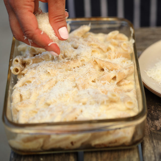Hand sprinkling grated cheese over gluten-free baked penne in a glass casserole dish on a wooden table