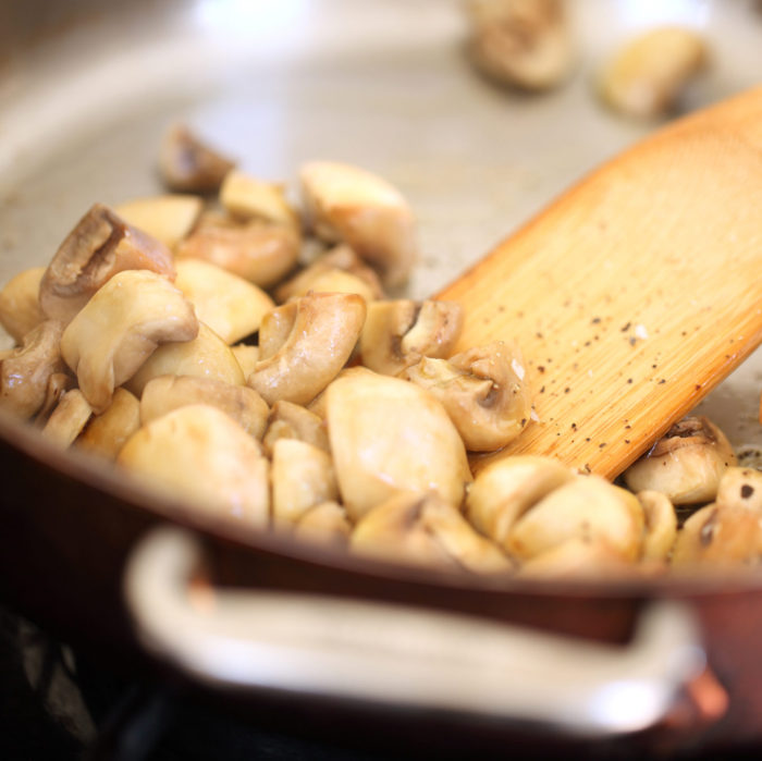 Sliced mushrooms sauteing in a skillet with a wooden spatula for beef stroganoff