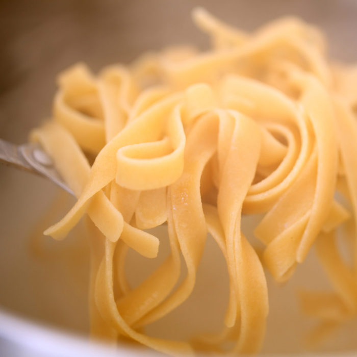 Fork-twirled brown rice tagliatelle close-up showing ribbon-like strands and tender texture in a blurred pot