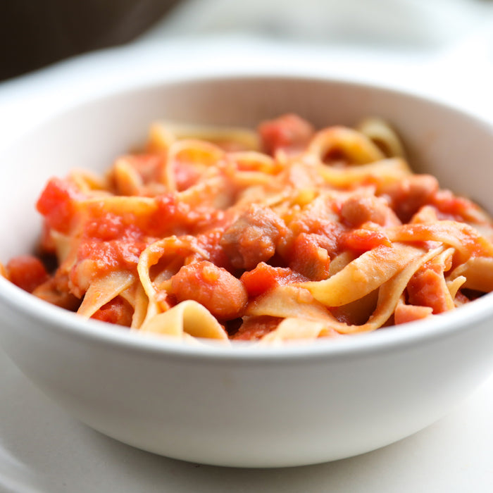 Close-up of gluten-free tagliatelle with borlotti beans in tomato sauce served in a white bowl, soft blurred background