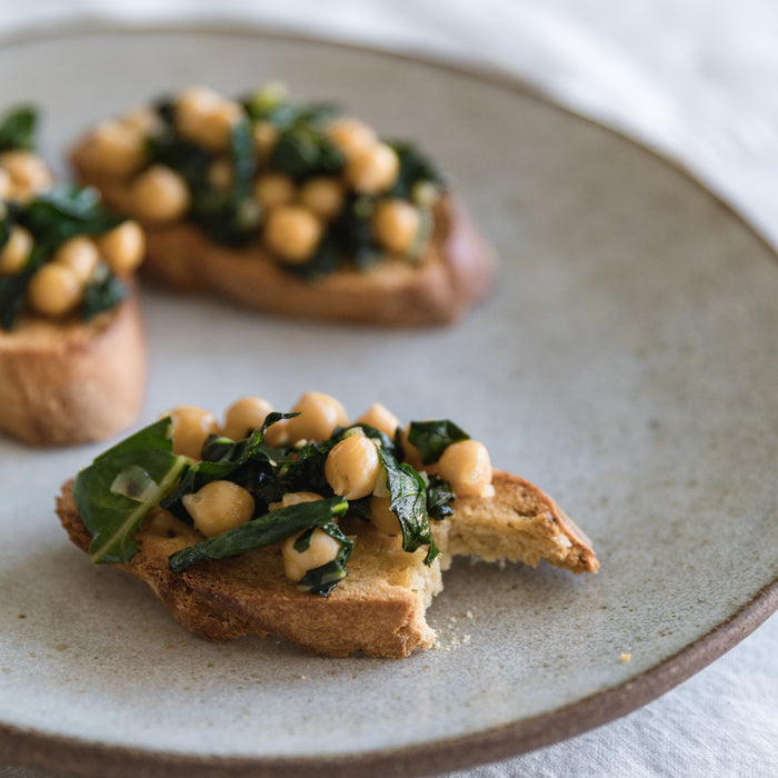 Sautéed chickpeas and wilted greens piled on a slice of toasted bread with a bite taken, on a ceramic plate