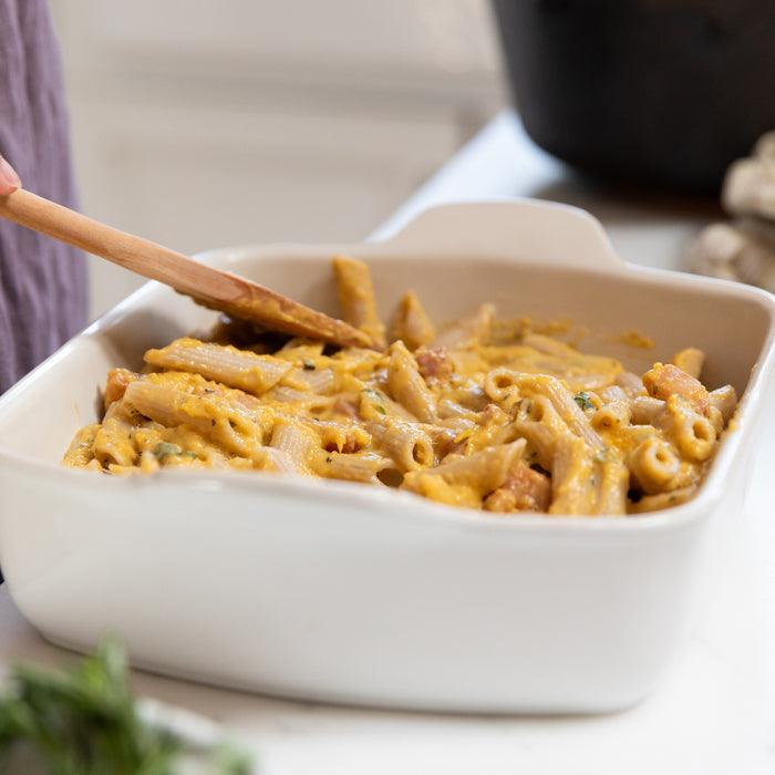 Gluten-free butternut squash penne al forno being stirred in a white baking dish in a kitchen