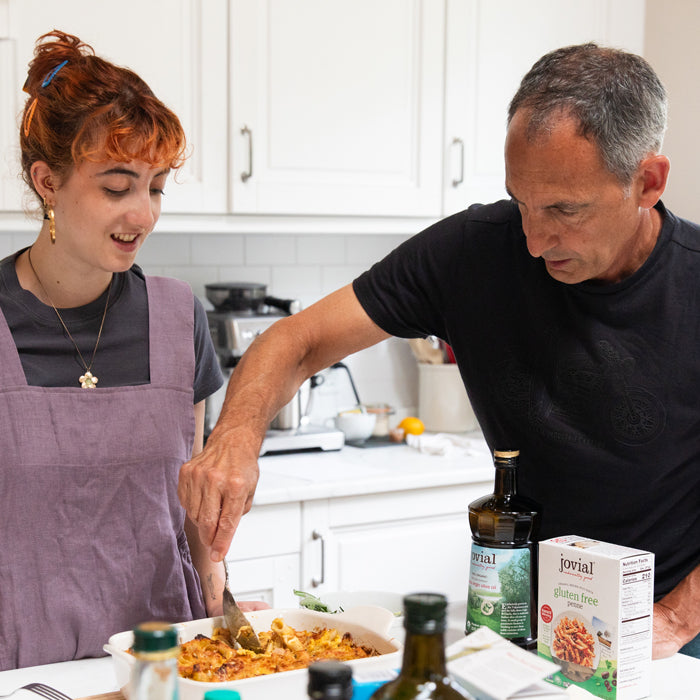 Young woman and man serving baked butternut squash penne from a casserole in a bright home kitchen