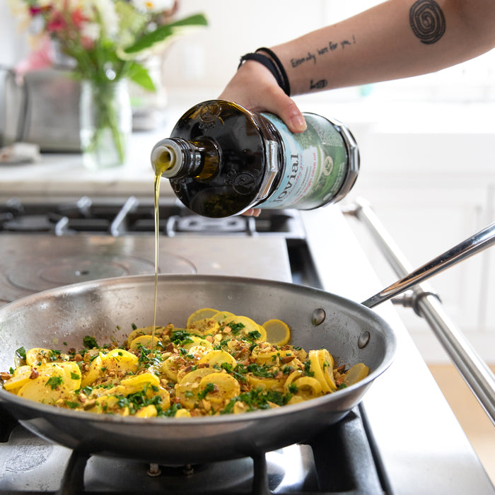 Hand pouring Jovial extra virgin olive oil over sautéed summer squash with chopped pistachios and parsley in a skillet