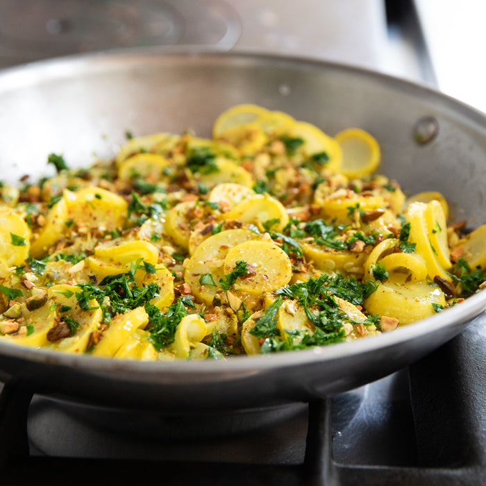 Sauteed sliced yellow summer squash with chopped pistachios and parsley in a stainless steel skillet on the stove