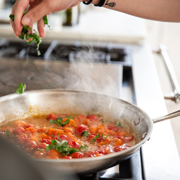 Chef sprinkling chopped basil over simmering cherry tomato sauce in a stainless-steel skillet on a stove, for tagliatelle pasta