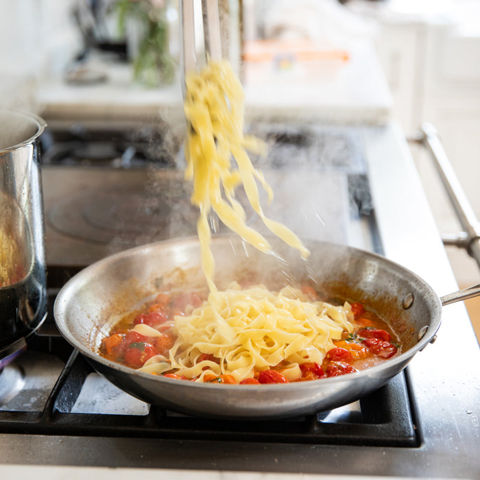 Tagliatelle being added to a skillet of cherry tomatoes on a stovetop, steam rising
