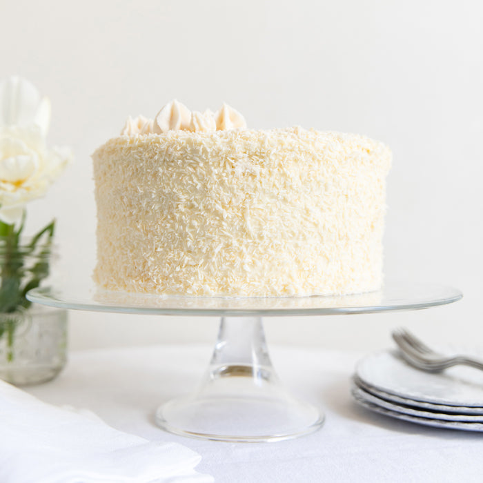 Front view of Einkorn coconut layer cake with shredded coconut coating and piped frosting on a glass stand