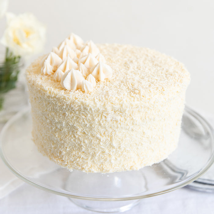 Three-quarter view of Einkorn coconut layer cake with shredded coconut coating and frosting dollops on a glass cake stand