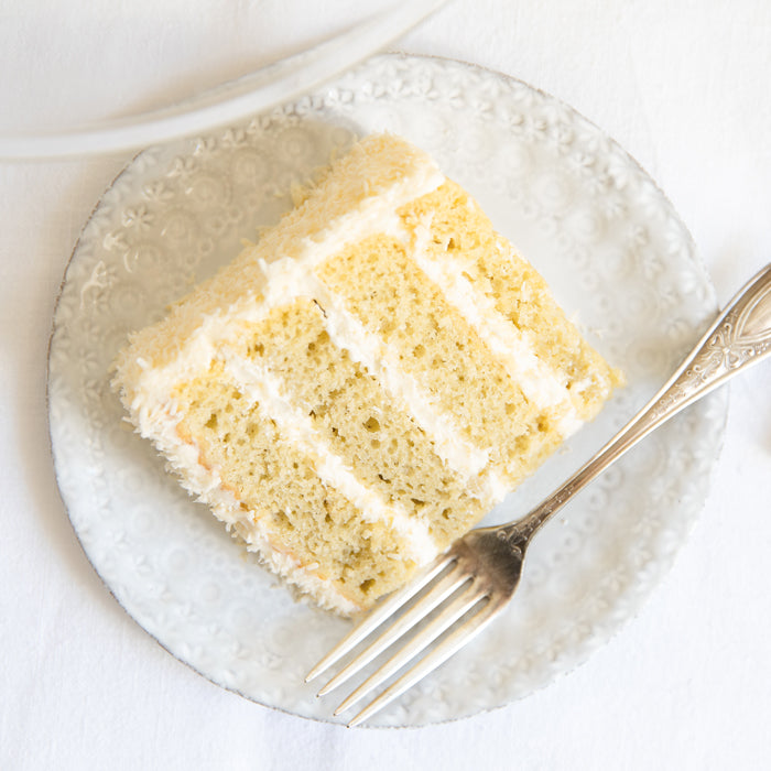 Top-down view of three-layer Einkorn coconut cake slice with shredded coconut and white frosting on a white plate with fork