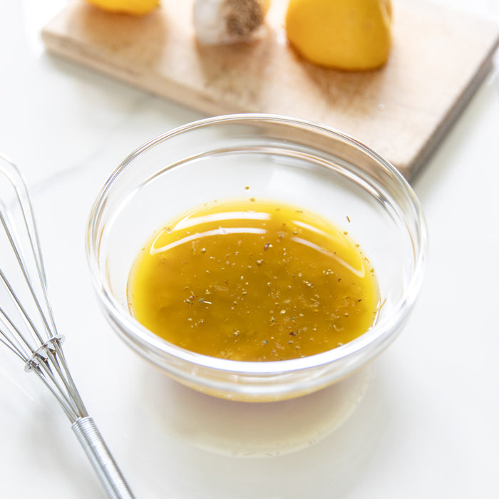Glass bowl of herbed olive oil vinaigrette with cracked pepper next to a whisk and halved lemon on a white countertop