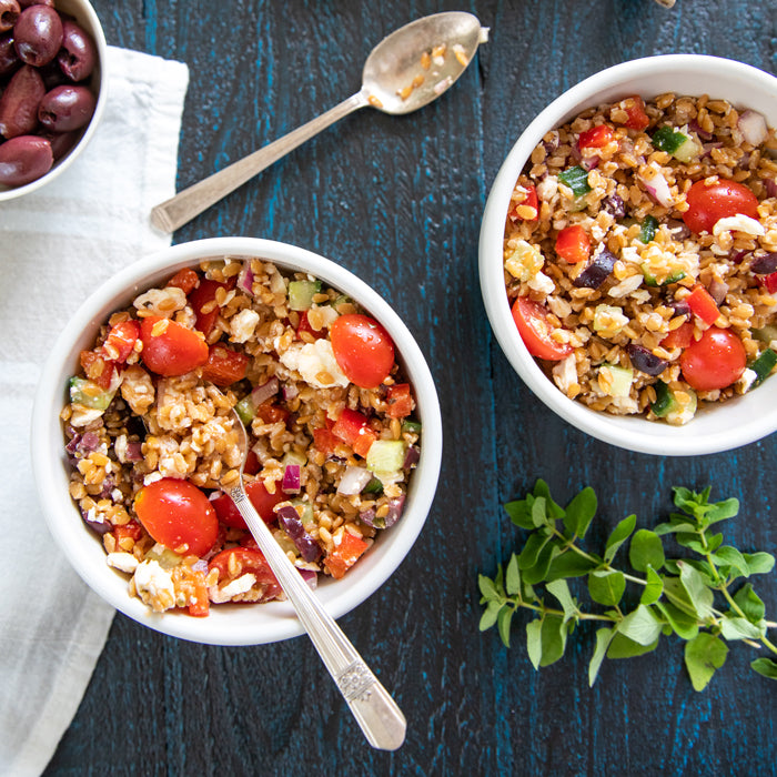 Two bowls of einkorn wheat berry salad served with cherry tomatoes, feta, olives, cucumber, and red onion on a dark wood surface
