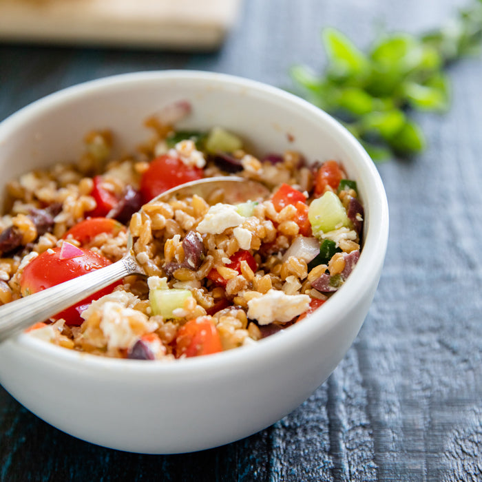 Close-up view of Greek Einkorn wheat berry salad showcasing chewy wheat berries, cherry tomatoes, cucumber, olives and feta in a white bowl on dark wood