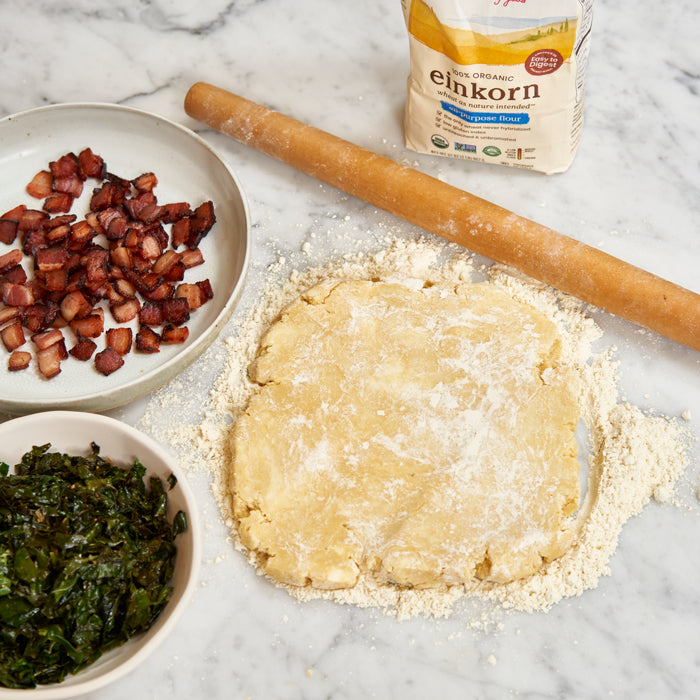 Rolled galette dough dusted with flour beside bowls of cooked bacon and sauteed kale, rolling pin, and bag of Einkorn flour