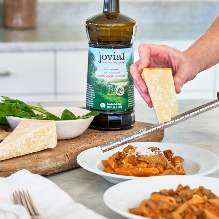 Hand grating Parmesan over pasta with a bottle of Jovial organic extra virgin olive oil on a kitchen counter