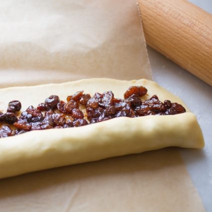 Einkorn rugelach dough being rolled around raisin filling on parchment with a rolling pin nearby