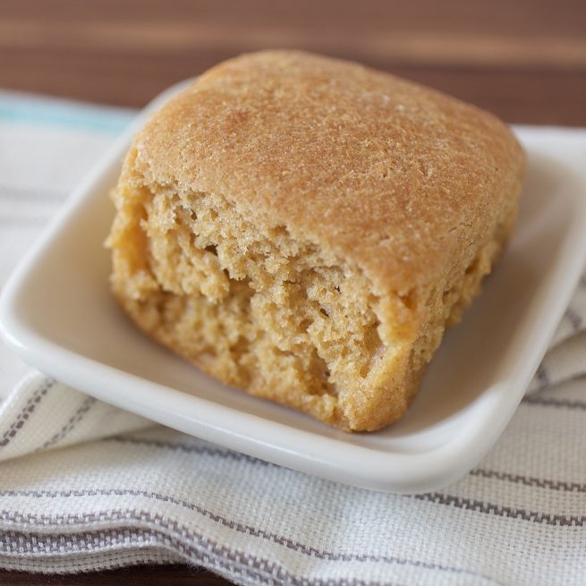Close-up front view of a whole grain einkorn dinner roll with torn interior showing soft crumb on a white dish over a striped cloth