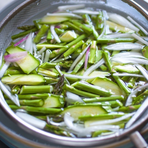 Asparagus, zucchini, and sliced red onion soaking in cold water in a metal strainer to remove dirt