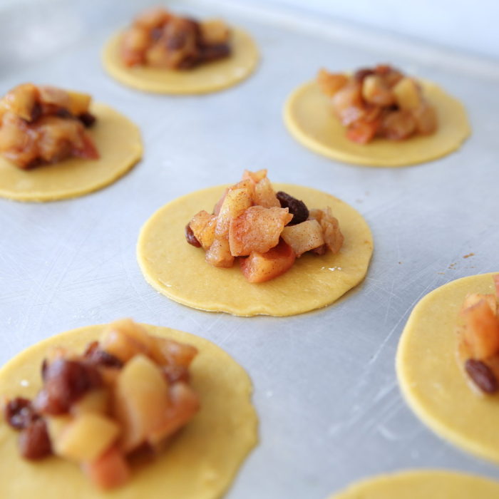 Einkorn apple hand pies being assembled on a baking sheet, round dough discs topped with spiced apple and raisin filling
