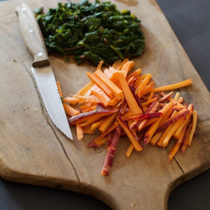 Julienned organic heirloom carrots and chopped rainbow chard on a wooden cutting board with a serrated knife