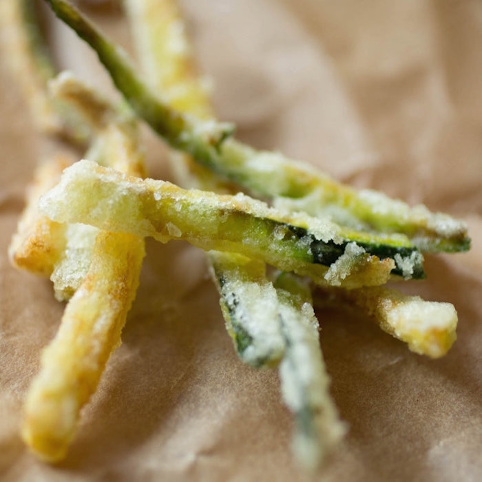 Close-up of Einkorn-battered fried zucchini sticks showing a light golden, crispy coating on brown parchment paper
