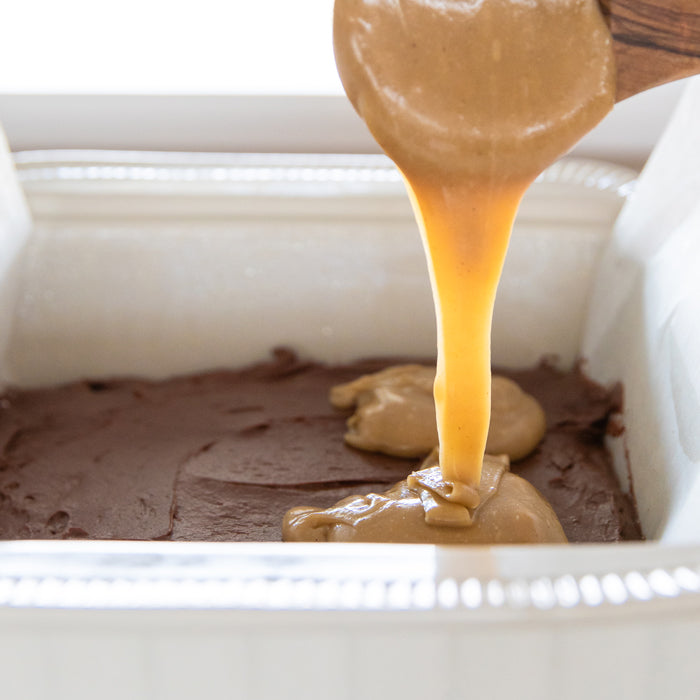 Halva being spooned and drizzled over fudgy einkorn brownie batter in a parchment-lined baking pan
