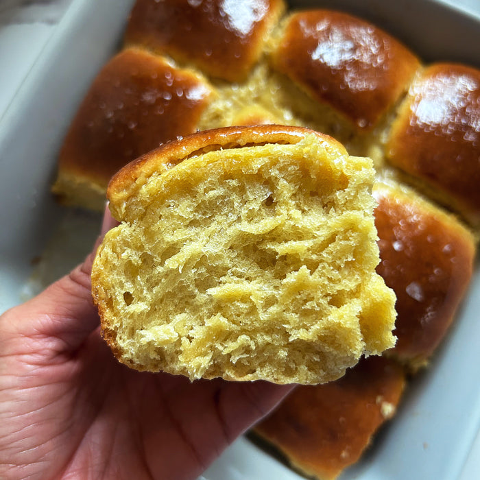 Close up of torn sourdough Hawaiian roll showing fluffy buttery yellow interior with glazed rolls in baking dish background