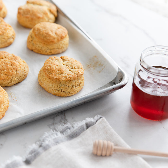 Three-quarter view of Einkorn honey biscuits on a baking sheet, golden-brown and flaky, with a jar of honey nearby