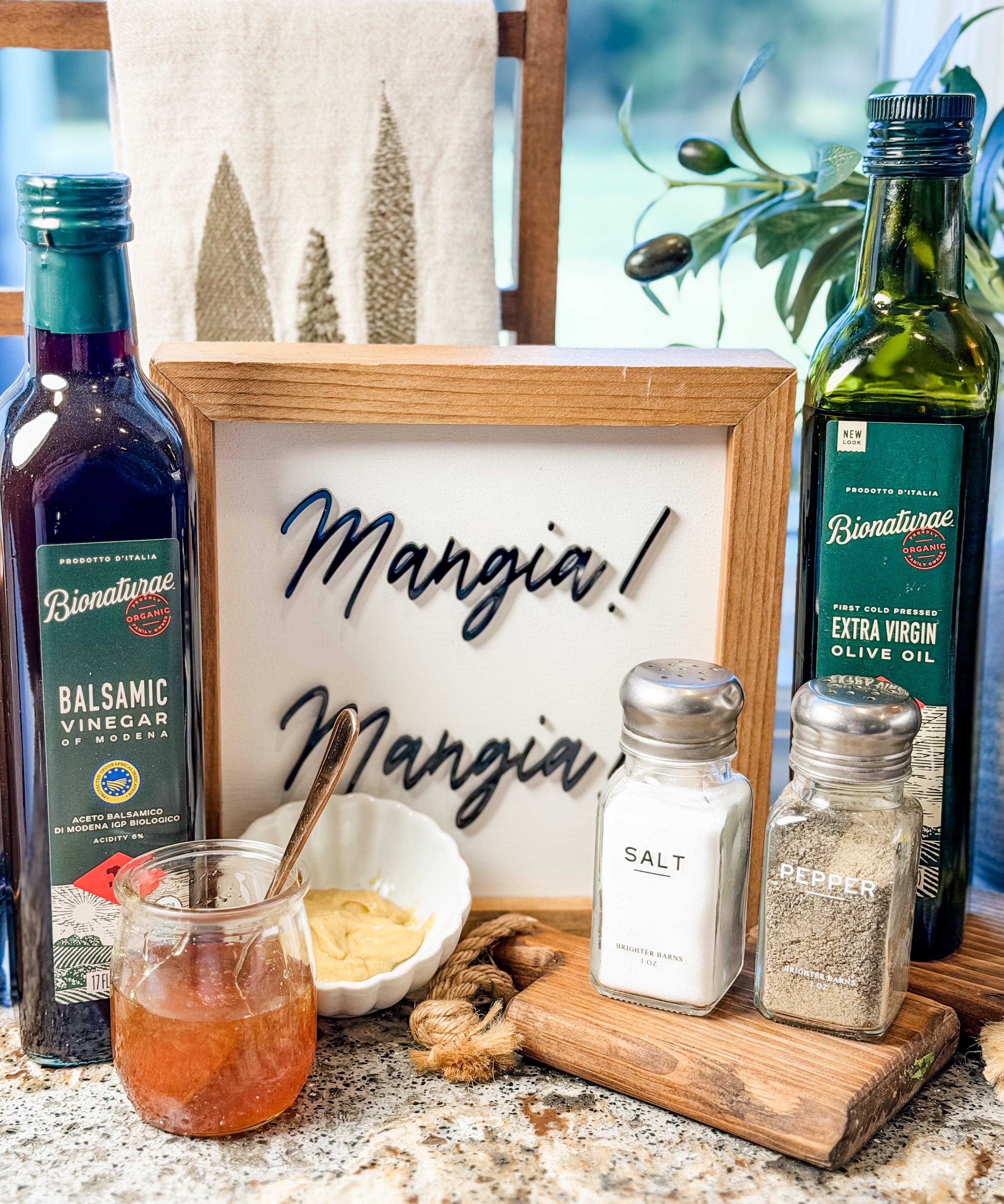 Bionaturae balsamic vinegar and extra virgin olive oil with salt and pepper on a kitchen counter before a framed Mangia sign