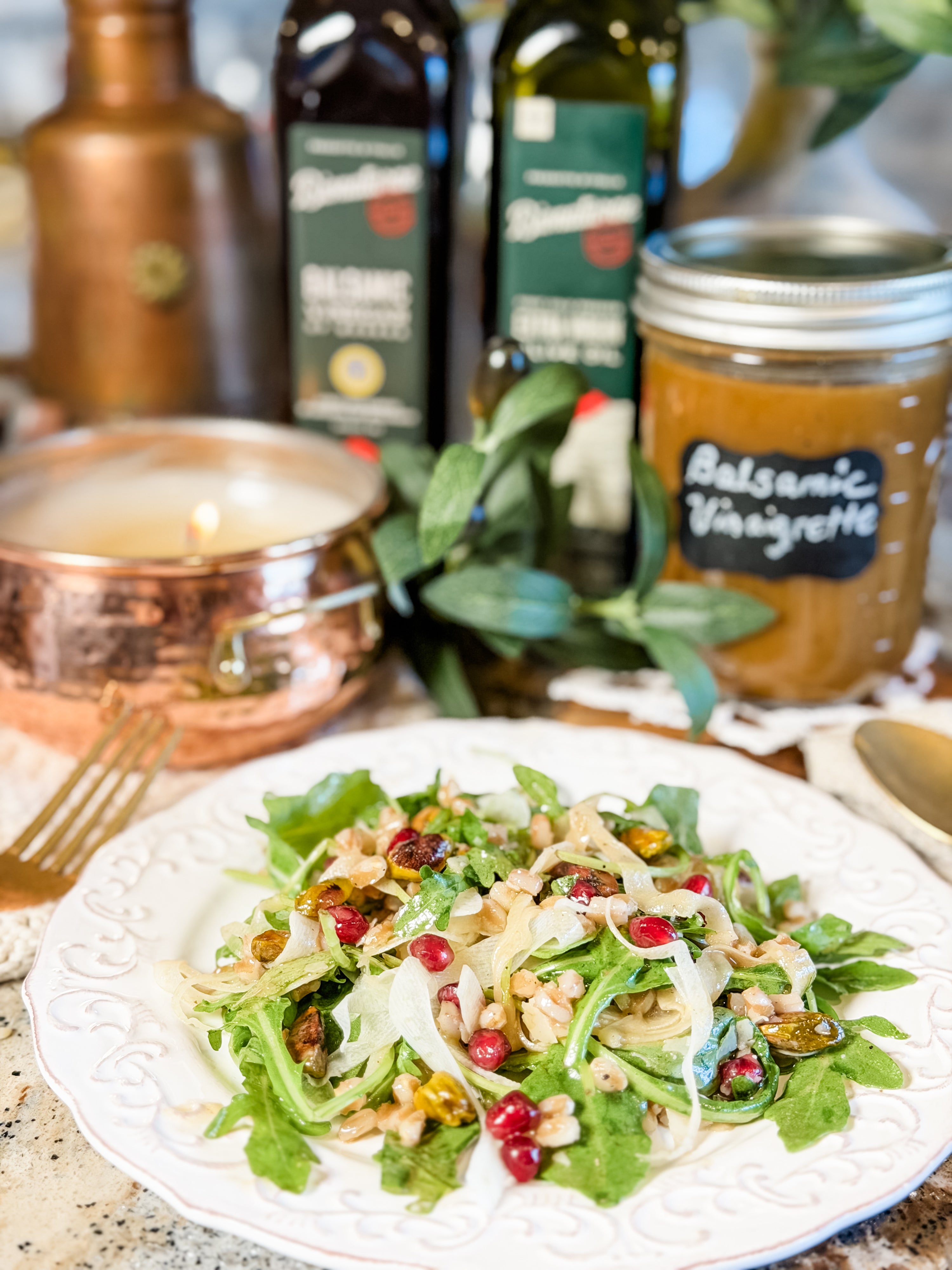 Salad of arugula, grains, pomegranate and pistachios with a jar labeled ‘Balsamic Vinaigrette’ and olive oil bottles behind