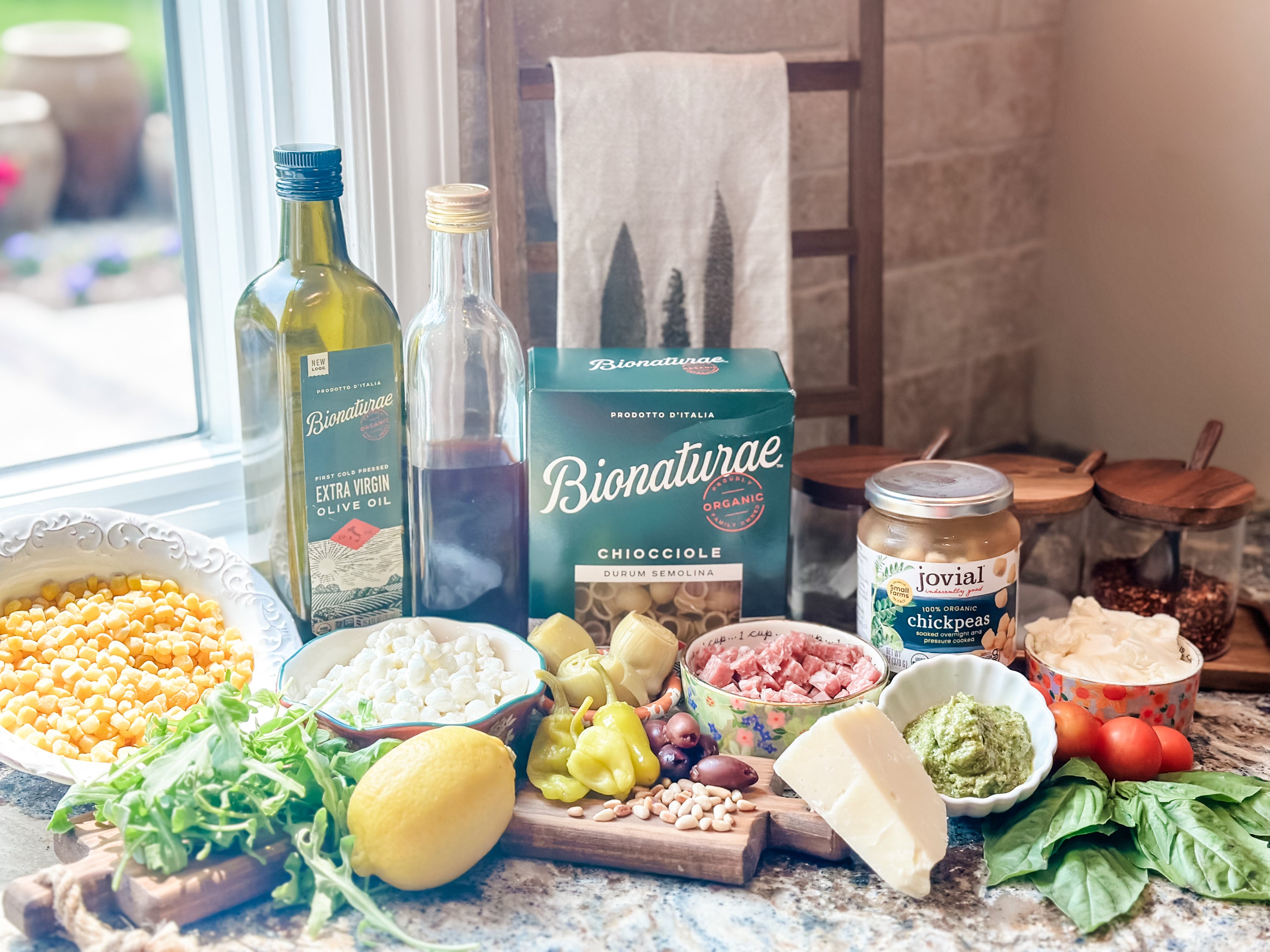 Kitchen counter with ingredients for a creamy pasta salad: Bionaturae pasta, Jovial chickpeas, olive oil, cheese, lemon