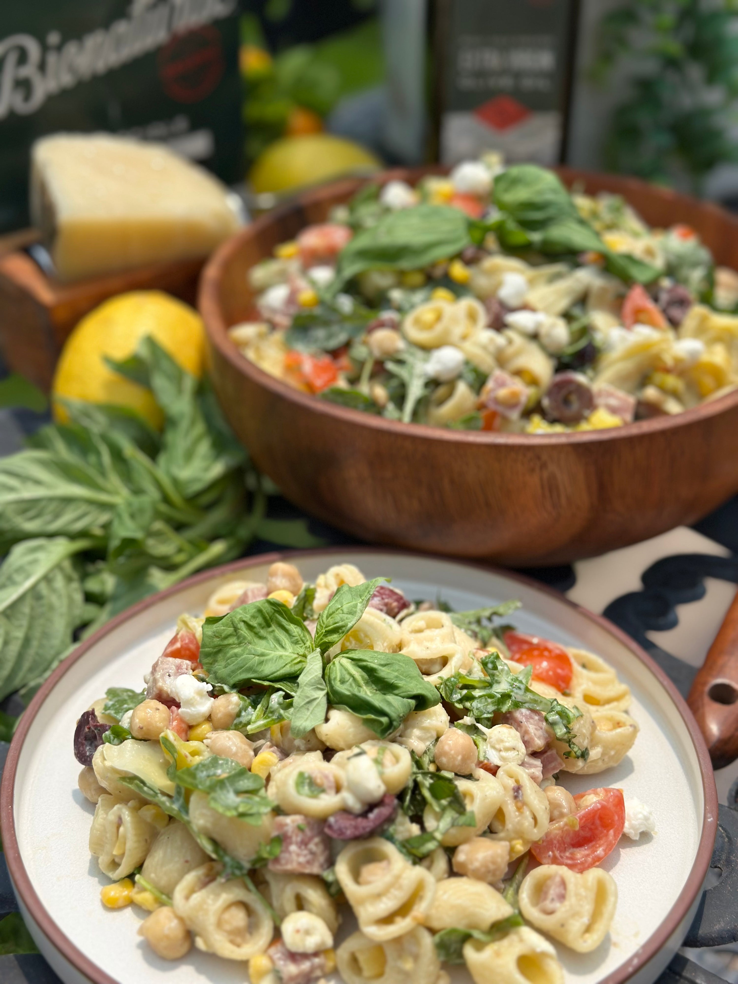 Plate and wooden bowl of creamy pasta salad without mayo, with basil, cherry tomatoes, olives, chickpeas, and ricotta