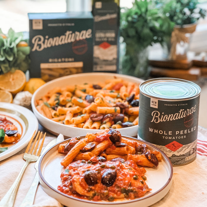 Plate of Italian penne in tomato, caper, and olive sauce beside a bowl of pasta and a Bionaturae whole peeled tomatoes can