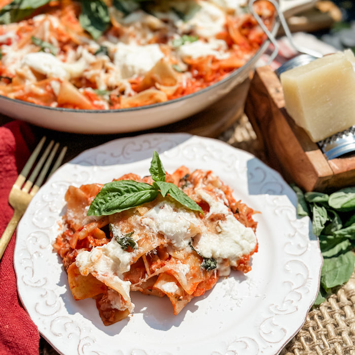 Italian skillet lasagna plated and garnished with ricotta and basil, skillet and cheese block on a rustic table outdoors