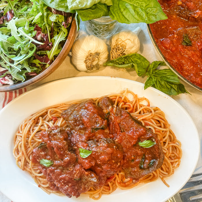 Steak pizzaiola plated over spaghetti with tomato sauce, basil leaves, garlic cloves, and a mixed green salad