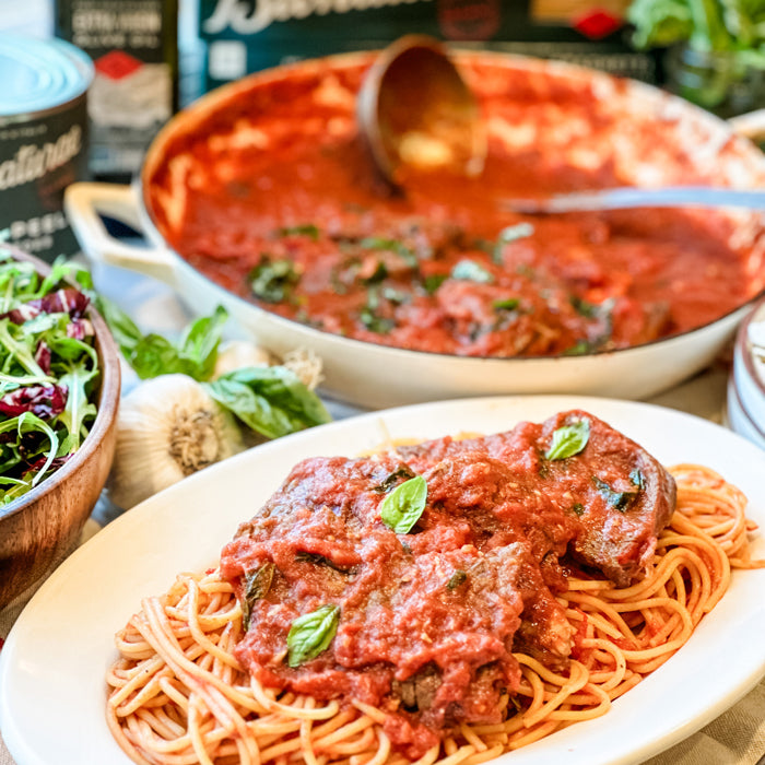 Steak pizzaiola atop spaghetti, topped with tomato sauce and basil, pan of sauce and a salad bowl in the background