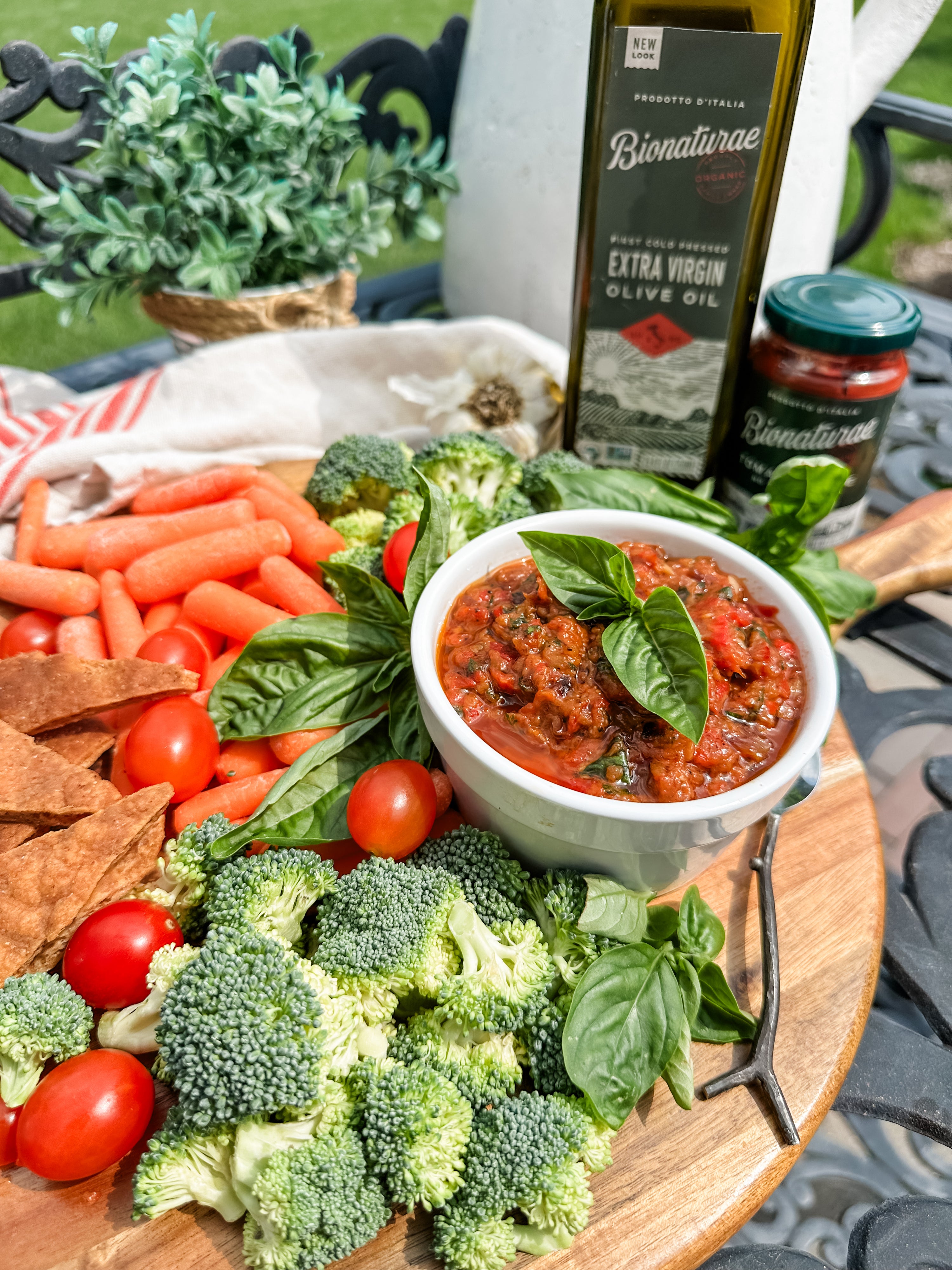 Roasted vegetable spread in a white bowl garnished with basil on a wooden board with broccoli, cherry tomatoes, baby carrots, pita chips and a Bionaturae olive oil bottle